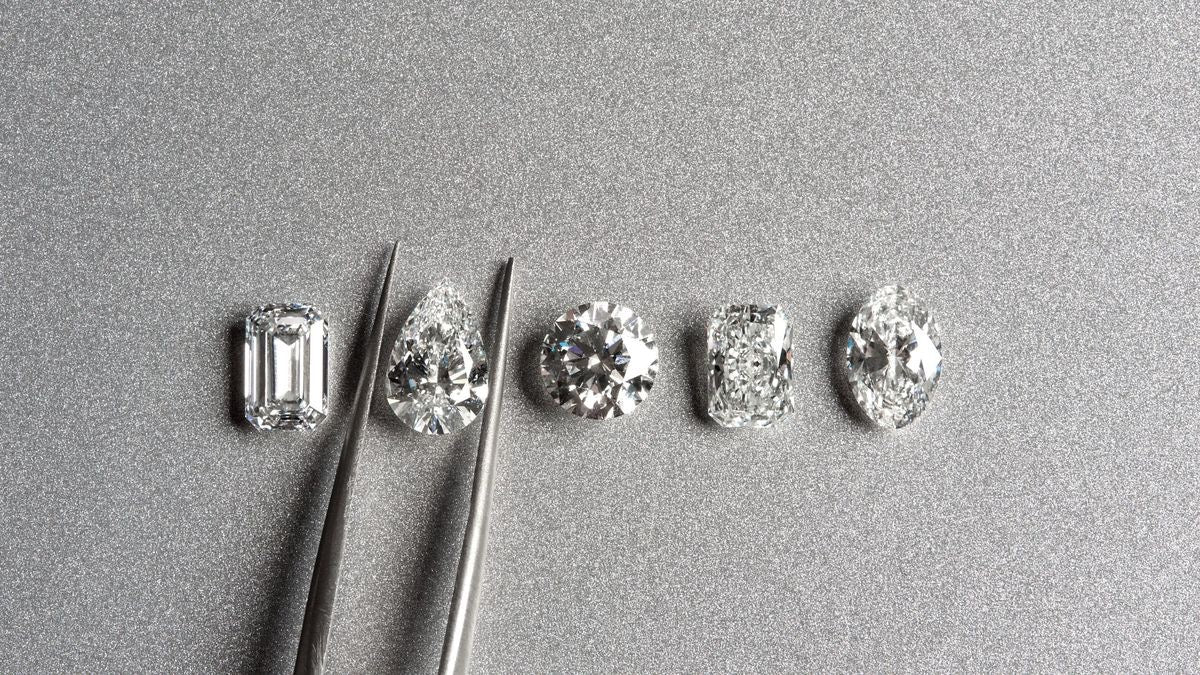 Five loose diamonds in a row: emerald, pear, round, radiant and oval cuts, with tweezers holding the pear
